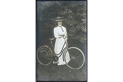 Kvinde iført hvid kjole og hat, står med cykel. Sted ukendt. Fotokort u/no. 
