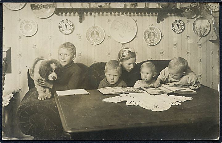 Børn kikker i bla. postkortalbum. På væggen forskellige platter bl.a. Aluminia Juleplatte 1906 og Aluminia  Livgardens Jubilæumsplatte 1658-1908. Fotokort u/no.