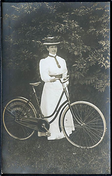 Kvinde iført hvid kjole og hat, står med cykel. Sted ukendt. Fotokort u/no. 