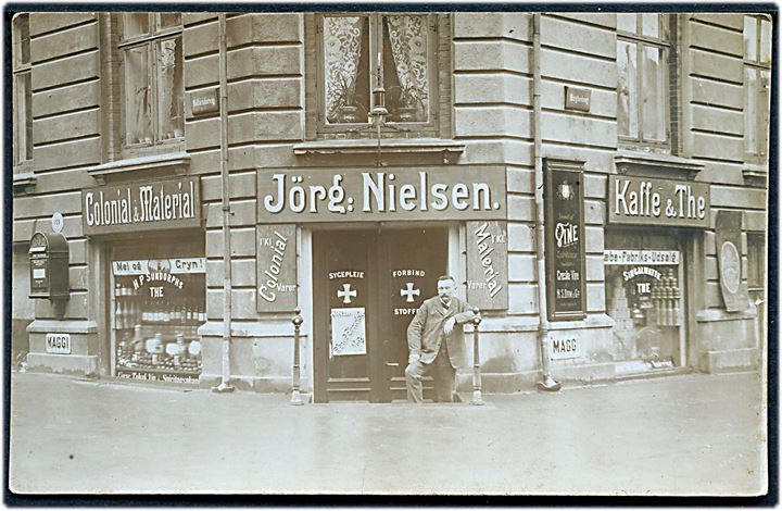 Enighedsvej hj. af Hollændervej med Jörg. Nielsen’s Colonial & Material, Kaffe & The handel. Fotokort u/no. Kvalitet 7