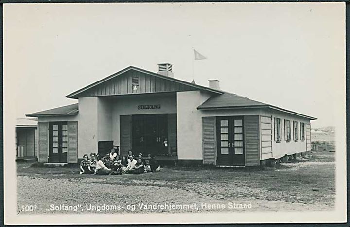 Ungdoms- og Vandrehjemmet Solfang, Henne Strand. En lille rundkreds med mennesker sidder udenfor og spiller guitar/synger. Fotokort no. 1007. 