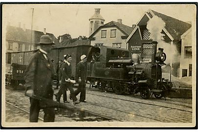 Godstog med mindre damplokomotiv i ukendt skandinavisk by med skilt Frederik Hansen Vine i baggrunden. Fotokort u/no.