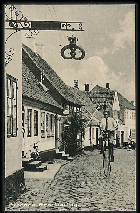Mand kommer cyklende på en væltepeter ved bageren, Brogade i Ærøskøbing. Hjørneknæk. Stenders no. 94848.