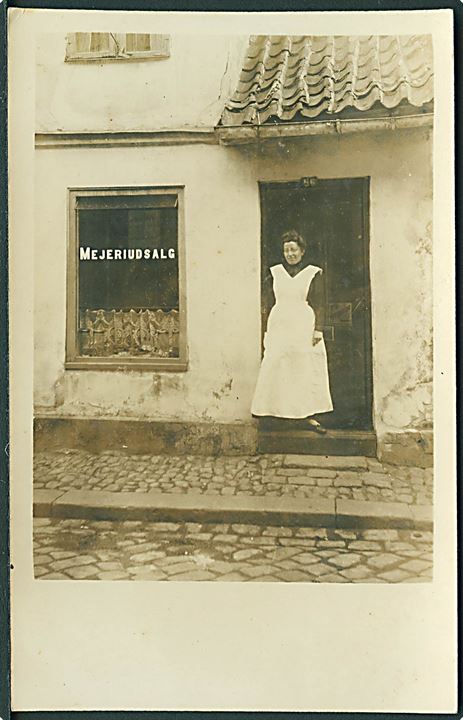 Facade no. 56. Mejeriudsalg. Kvinde står i døren. Sted ukendt. Fotokort u/no.